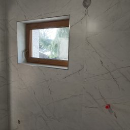 WLAD-BUD - Wykończona ściana w łazience z oknem w drewnianej ramie. Płytki imitujące marmur z widocznymi fugami. Otwór na instalację elektryczną. Częściowo widoczne narzędzia.