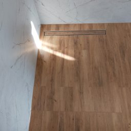 WLAD-BUD - Wykończona łazienka z drewnopodobną podłogą i odpływem liniowym. Ściany wyłożone płytkami imitującymi marmur. Widoczne fragmenty butów w dolnej części kadru.