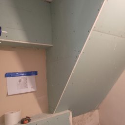 WLAD-BUD - Ściana poddasza z płyt karton-gips, widoczne łączenia i wkręty. Na ścianie plan z niebieską taśmą, obok rolka papieru i miarka. Etap prac wykończeniowych.