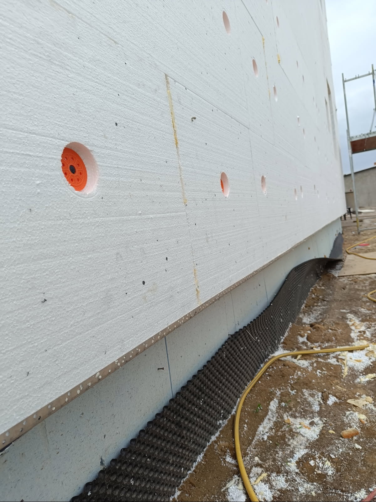 Ocieplenie budynku styropianem z widocznymi kołkami montażowymi i folią kubełkową przy fundamencie. Prace elewacyjne w toku, widok z dołu.