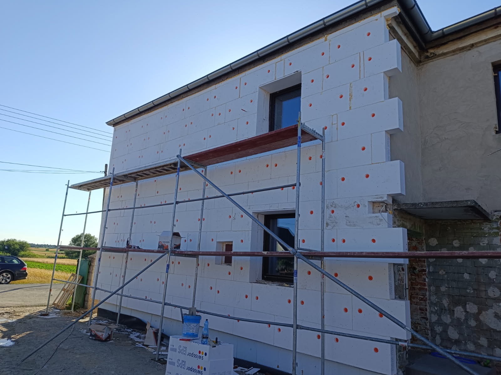 Ocieplanie elewacji styropianem z rusztowaniem w Poznaniu. Widoczne płyty Swissopor EPS z pomarańczowymi kołkami. Częściowo ocieplony budynek z oknami.