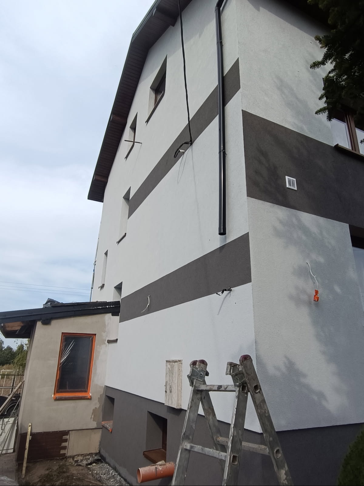 Dwukolorowa elewacja domu po remoncie, z widoczną drabiną i fragmentem dobudówki w trakcie prac. Szare pasy na białej fasadzie, rynna pionowa.