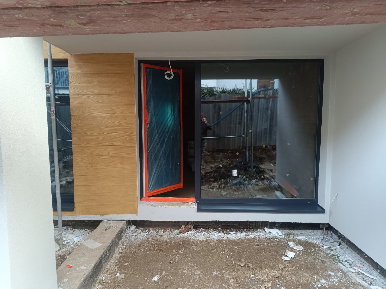 Nowoczesna elewacja z drewnopodobną okładziną i dużym oknem przesuwnym. Otwarta osłonięta folią drzwi balkonowe. Widoczne rusztowania w tle.