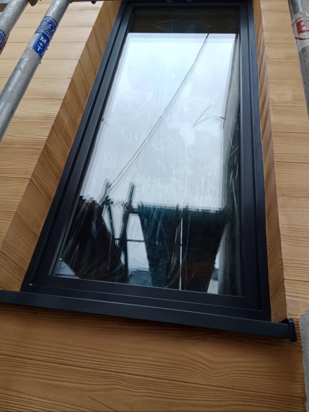 Nowe okno z czarną ramą, zamontowane w elewacji z drewnianą okładziną. Widoczne rusztowanie. Okno zabezpieczone folią ochronną.