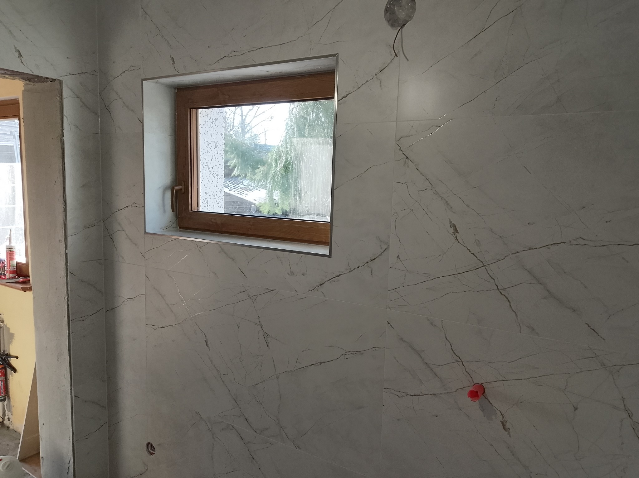 Wykończona ściana w łazience z oknem w drewnianej ramie. Płytki imitujące marmur z widocznymi fugami. Otwór na instalację elektryczną. Częściowo widoczne narzędzia.