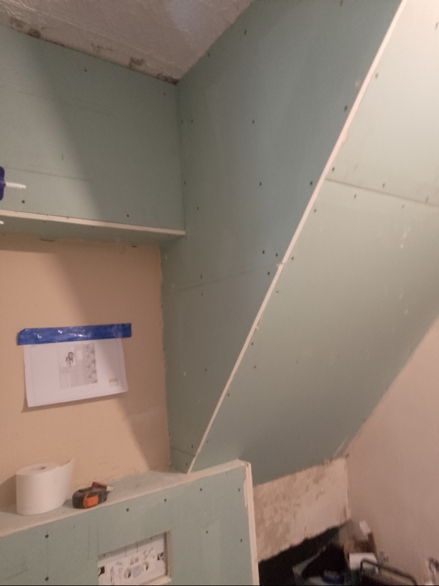 Ściana poddasza z płyt karton-gips, widoczne łączenia i wkręty. Na ścianie plan z niebieską taśmą, obok rolka papieru i miarka. Etap prac wykończeniowych.