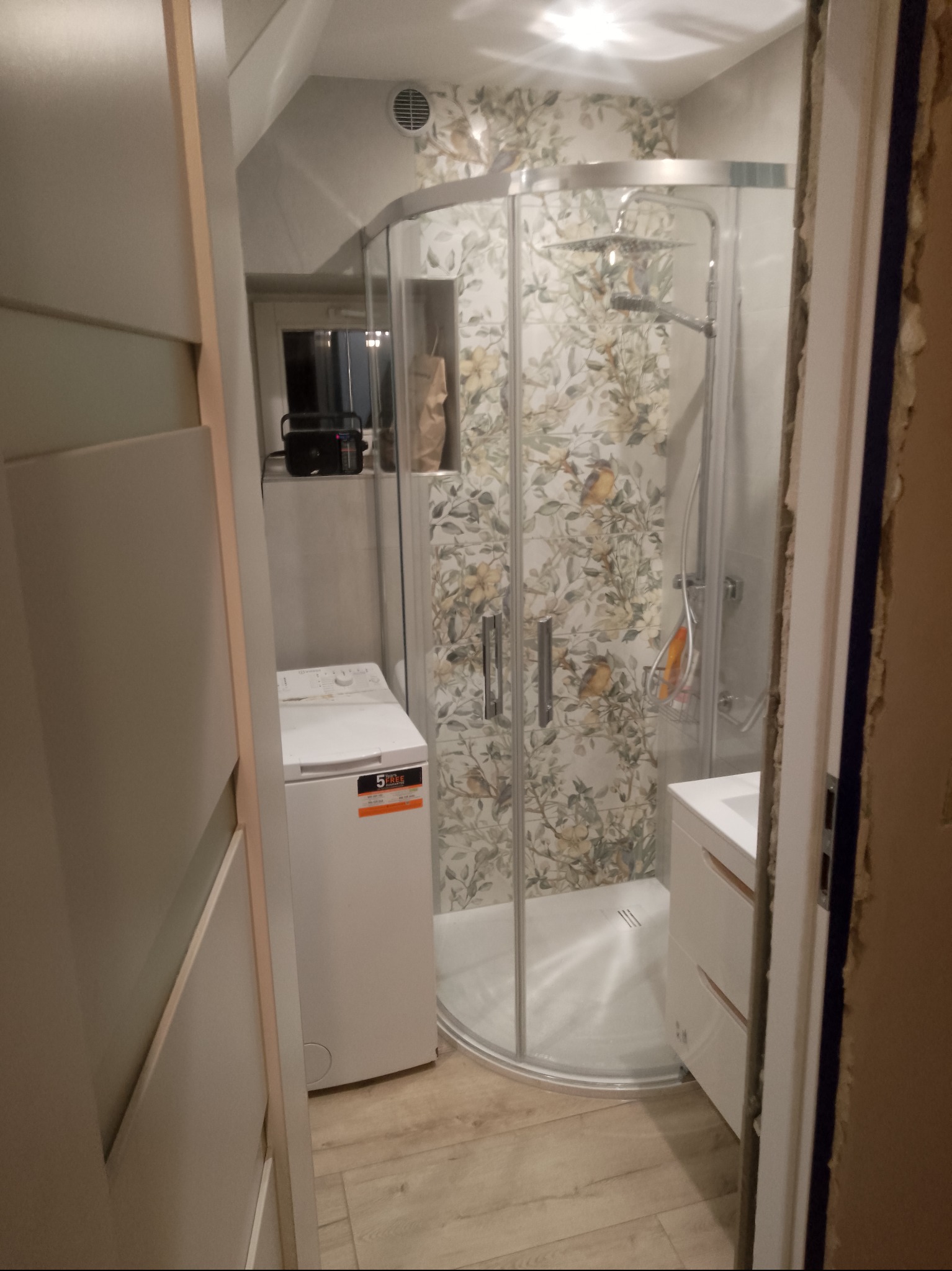 Kompaktowa łazienka po remoncie: kabina prysznicowa z wzorzystymi płytkami, pralka i umywalka z szafką. Widok zza framugi drzwi.