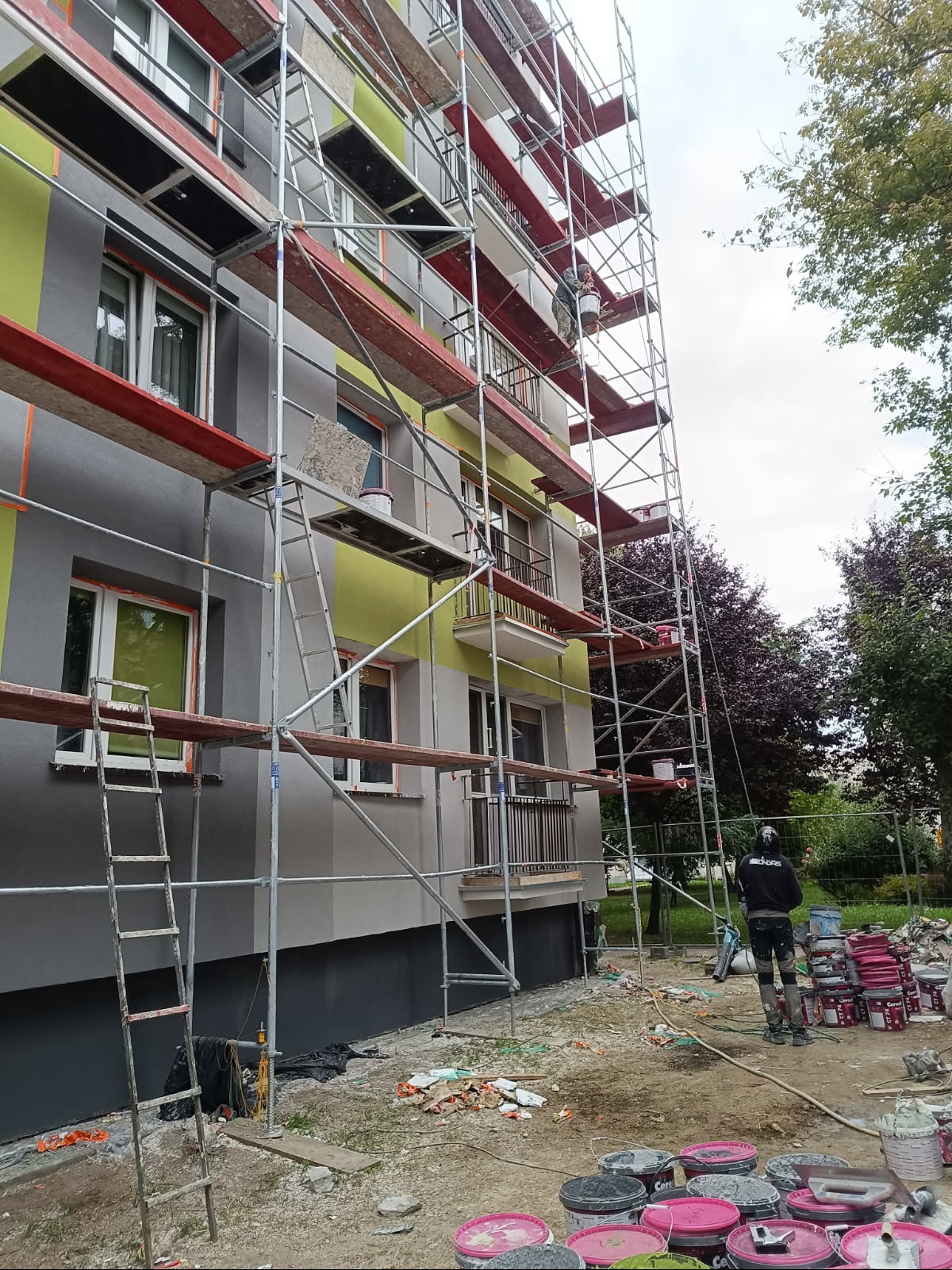 Malowanie elewacji bloku z rusztowania. Widoczne puszki z farbą, drabina i pracownik w trakcie pracy. Częściowo pomalowana ściana w odcieniach szarości i zieleni.