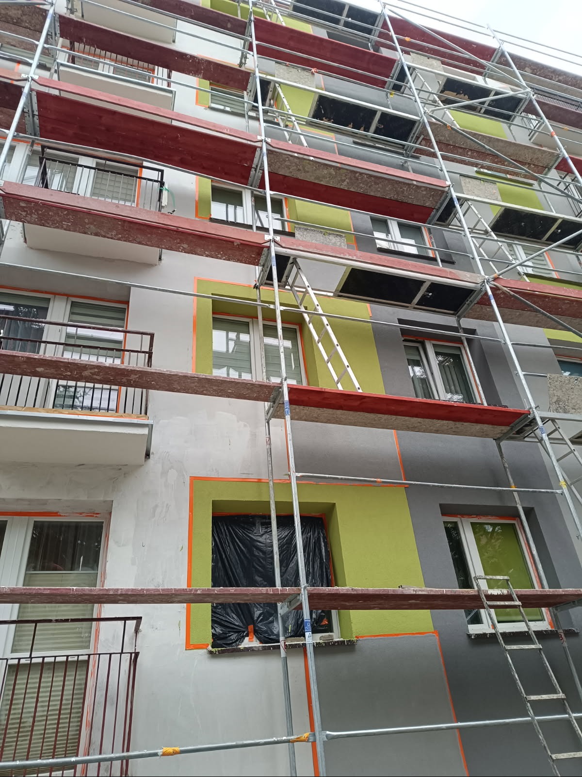 Budynek w trakcie malowania elewacji, widoczne rusztowania, okna zabezpieczone folią, fragmenty pomalowane na szaro i zielono, detale wykończeniowe.