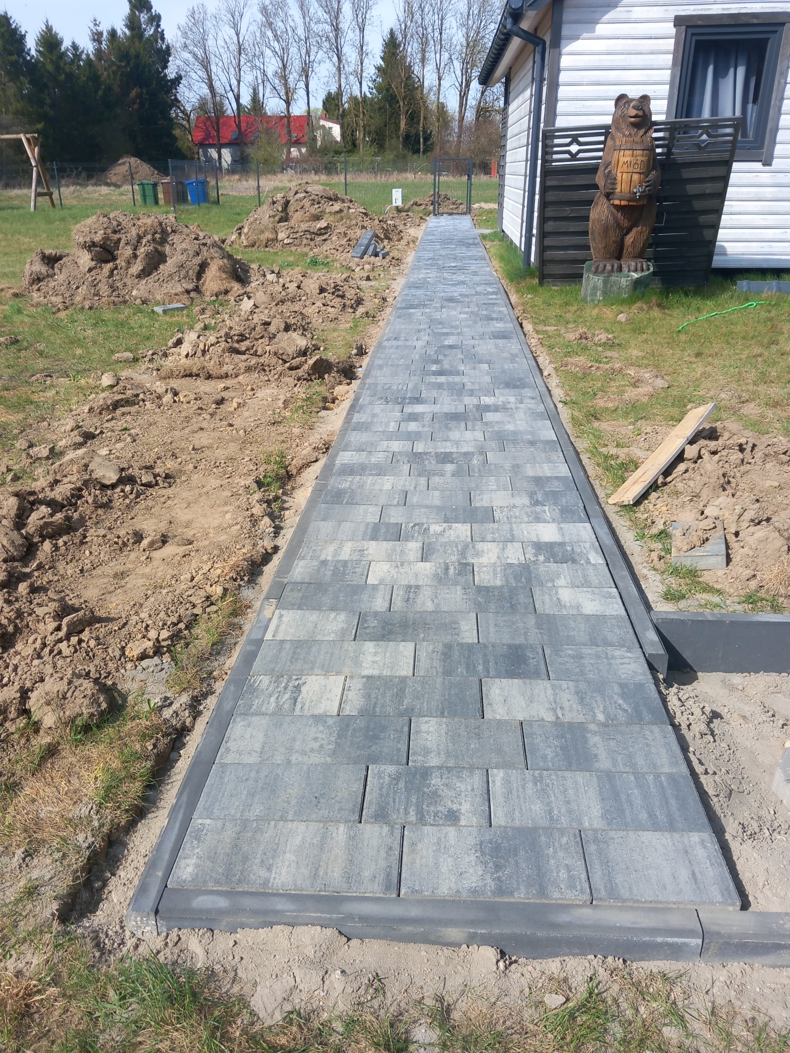 Ułożona, długa ścieżka z szarej kostki brukowej, obrzeża wykończone ciemniejszymi elementami. W tle dom z drewnianą rzeźbą niedźwiedzia i hałdy ziemi.