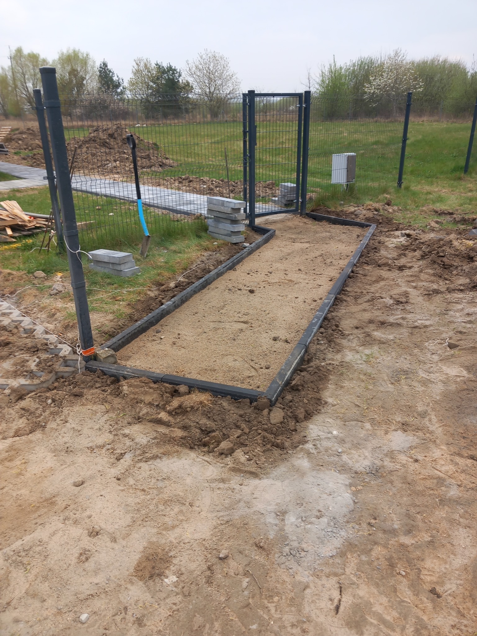 Przygotowany teren pod ścieżkę z obrzeżami, widoczny piasek, kostka brukowa i narzędzia. W tle siatka ogrodzeniowa i zieleń. Etap prac brukarskich.