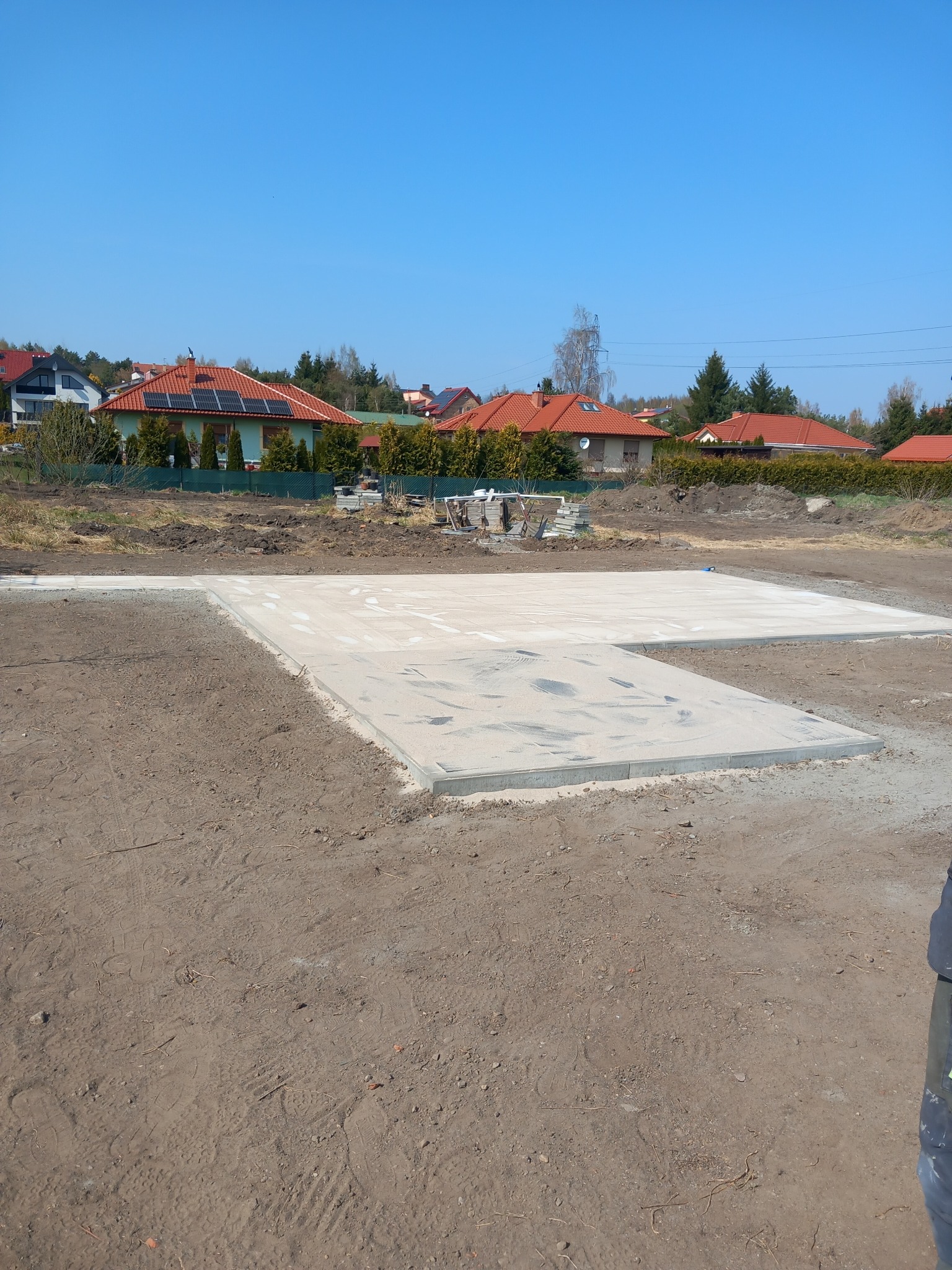 Betonowa podbudowa pod budynek na działce, widoczne domy w tle. Teren wyrównany, przygotowany pod dalsze prace budowlane. Słoneczny dzień, błękitne niebo.