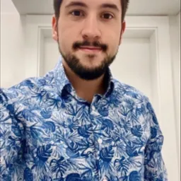 Portret uśmiechniętego mężczyzny w niebieskiej koszuli w tropikalne wzory, sfotografowanego w jasnym wnętrzu z białymi drzwiami i okrągłą lampą sufitową.