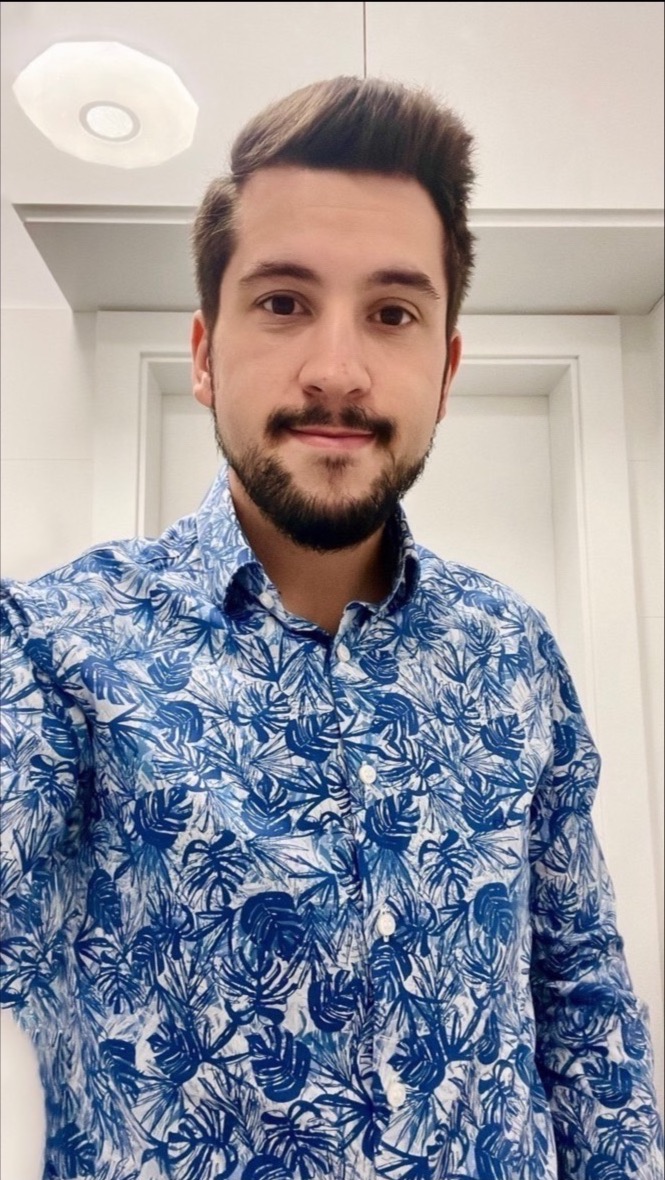 Portret uśmiechniętego mężczyzny w niebieskiej koszuli w tropikalne wzory, sfotografowanego w jasnym wnętrzu z białymi drzwiami i okrągłą lampą sufitową.