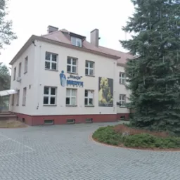 Dwupiętrowy budynek z beżową elewacją, ciemnoczerwonym cokołem, białymi oknami, logo 'Stacja Medyk Otwock' i portretem na ścianie, otoczony kostką brukową i drzewami iglastymi.