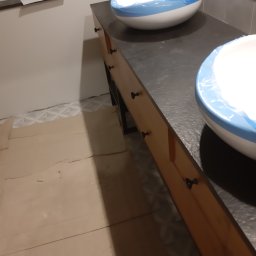 Praus Piotr Kowalczyk - Dwie białe umywalki z niebieskimi brzegami, zabezpieczone folią, stoją na drewnianej szafce z czarnym blatem i małymi czarnymi uchwytami, podłoga zabezpieczona kartonem.