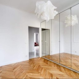 Praus Piotr Kowalczyk - Jasne wnętrze pokoju z parkietem ułożonym w jodełkę, białymi ścianami z dekoracyjnymi listwami, lustrzaną szafą w zabudowie i ozdobną lampą wiszącą.