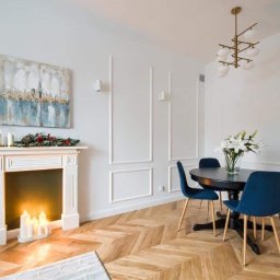 Praus Piotr Kowalczyk - Jasne wnętrze z kominkiem, nad którym wisi abstrakcyjny obraz; obok stół z ciemnym blatem i niebieskimi krzesłami na parkiecie ułożonym w jodełkę; w tle dekoracyjne listwy na ścianie