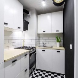 Praus Piotr Kowalczyk - Kompaktowa kuchnia w stylu retro z białą zabudową, drewnianym blatem, czarno-białą podłogą w szachownicę i dekoracyjnym zegarem ściennym.