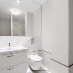 Praus Piotr Kowalczyk - Nowoczesna, jasna łazienka z białymi marmurowymi płytkami, wiszącą toaletą i szafką umywalkową z złotymi uchwytami, odbicie kabiny prysznicowej w lustrze.