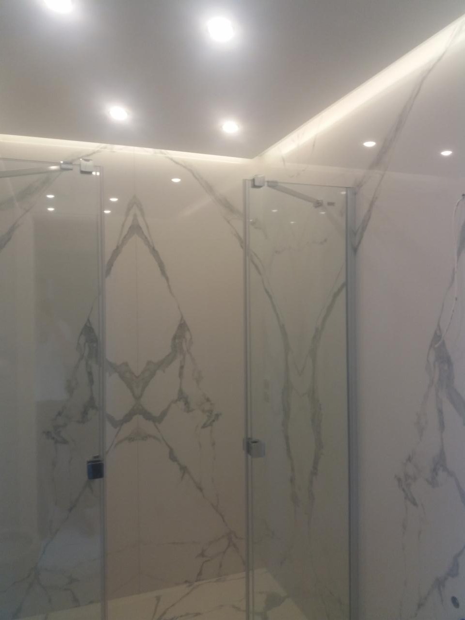 Kabina prysznicowa z dwoma szklanymi drzwiami z chromowanymi zawiasami, wykończona płytkami imitującymi marmur z szarymi żyłami, oświetlenie punktowe w suficie