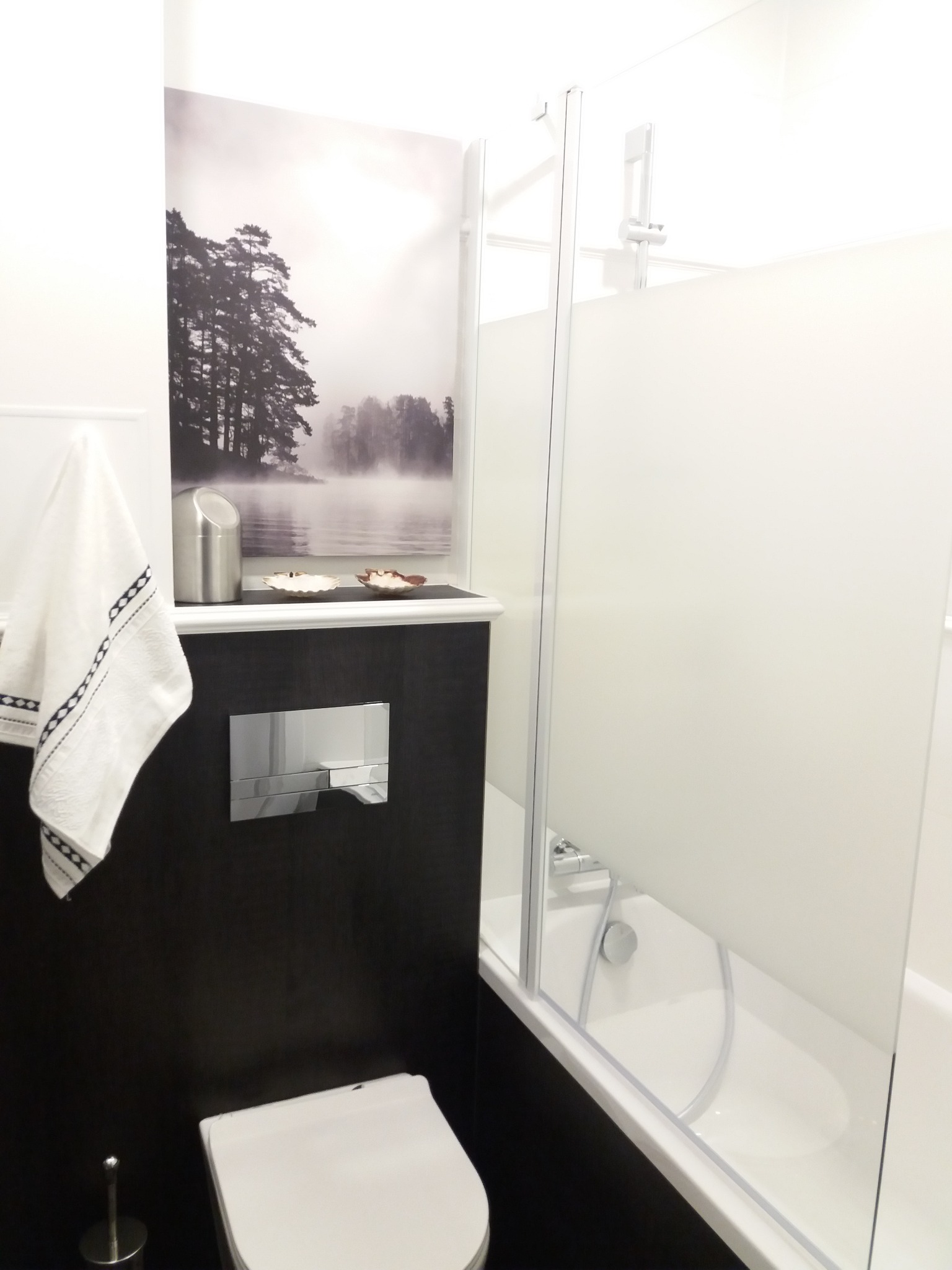 Nowoczesna łazienka z czarną ścianą, wiszącą toaletą, szklanym parawanem nawannowym i dekoracyjnym obrazem z motywem leśnym nad spłuczką.