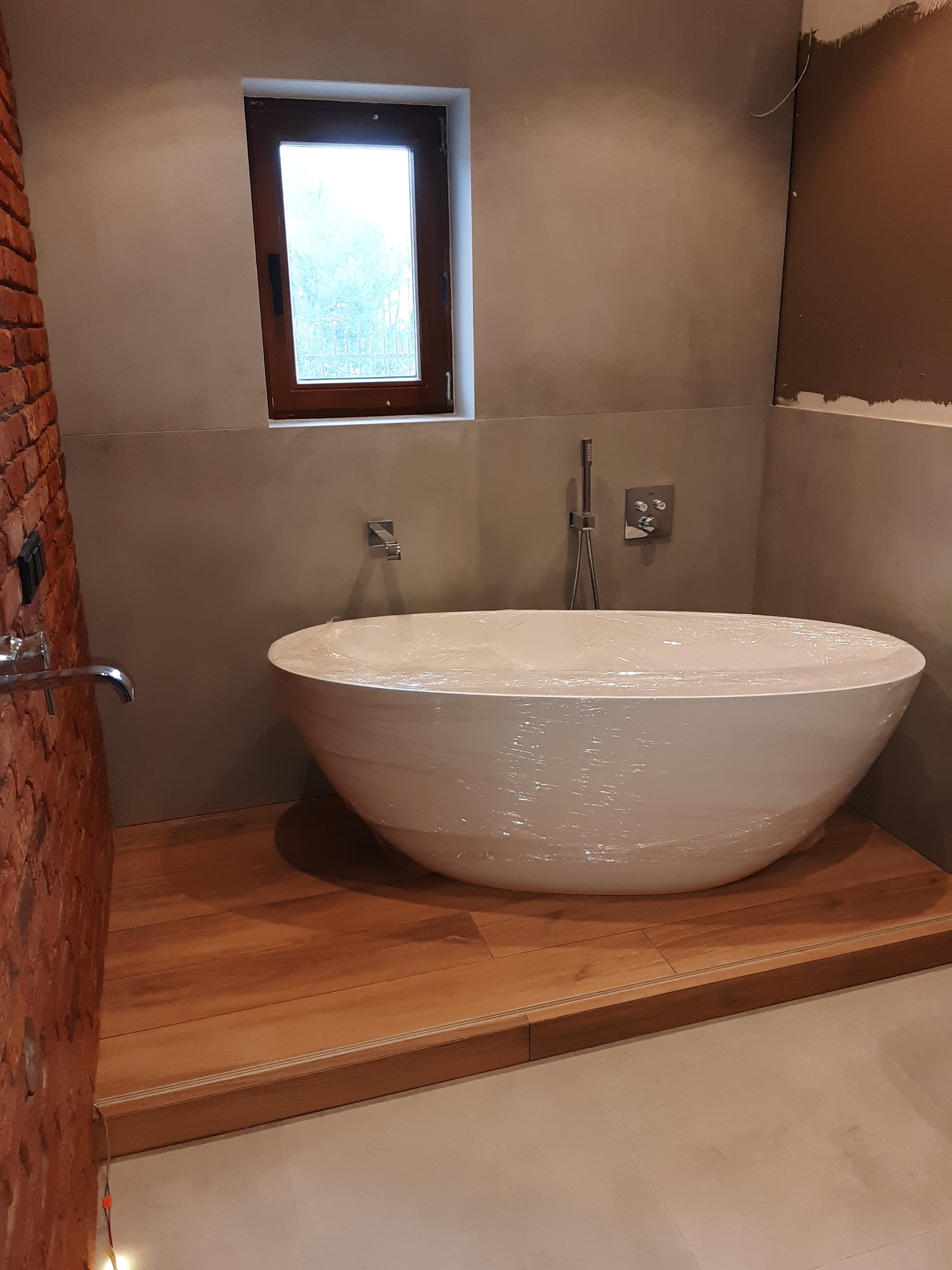 Nowa owalna wanna wolnostojąca, jeszcze w folii ochronnej, umieszczona na drewnianym podeście w surowej łazience z ceglanym akcentem i małym oknem.