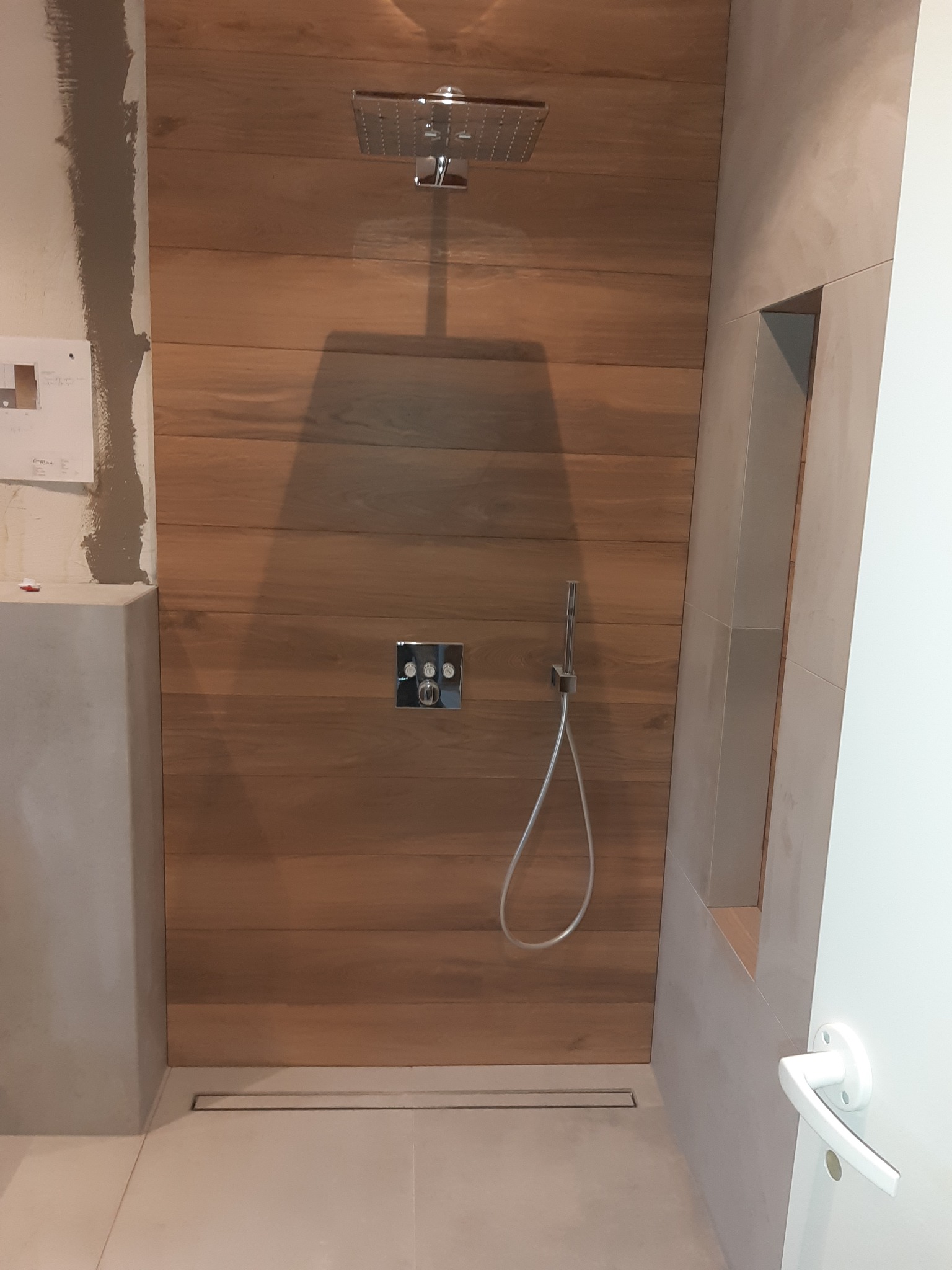 Nowoczesna kabina prysznicowa z drewnopodobną okładziną, deszczownicą i odpływem liniowym w podłodze, częściowo wykończona, z widocznymi śladami prac na ścianie obok.