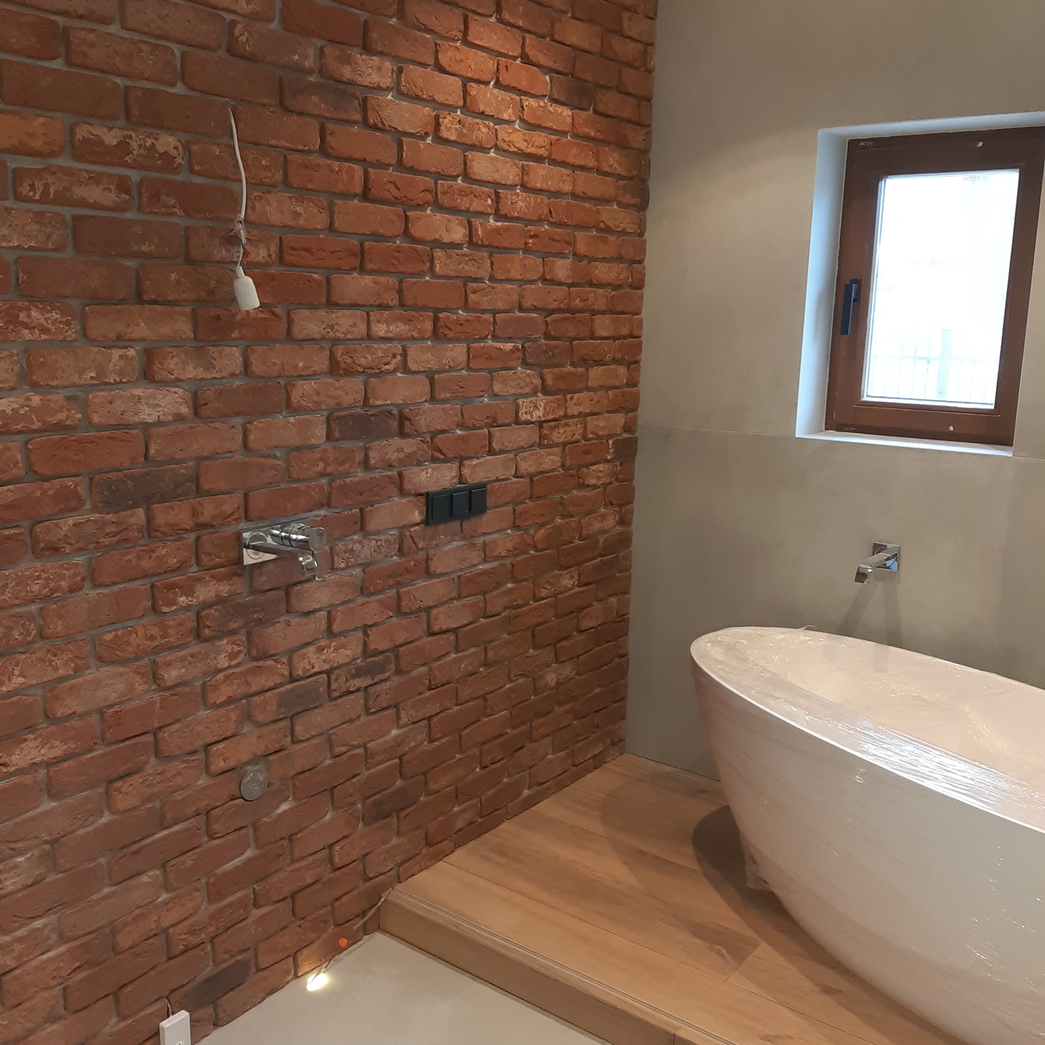 Łazienka w trakcie wykańczania: ściana z czerwonej cegły z chromowaną baterią i włącznikami, wolnostojąca wanna owinięta folią ochronną, drewniana podłoga, okno w drewnianej ramie.