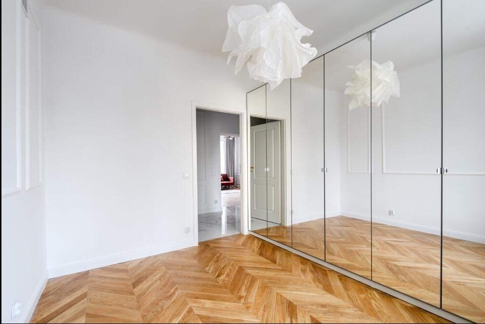 Jasne wnętrze pokoju z parkietem ułożonym w jodełkę, białymi ścianami z dekoracyjnymi listwami, lustrzaną szafą w zabudowie i ozdobną lampą wiszącą.