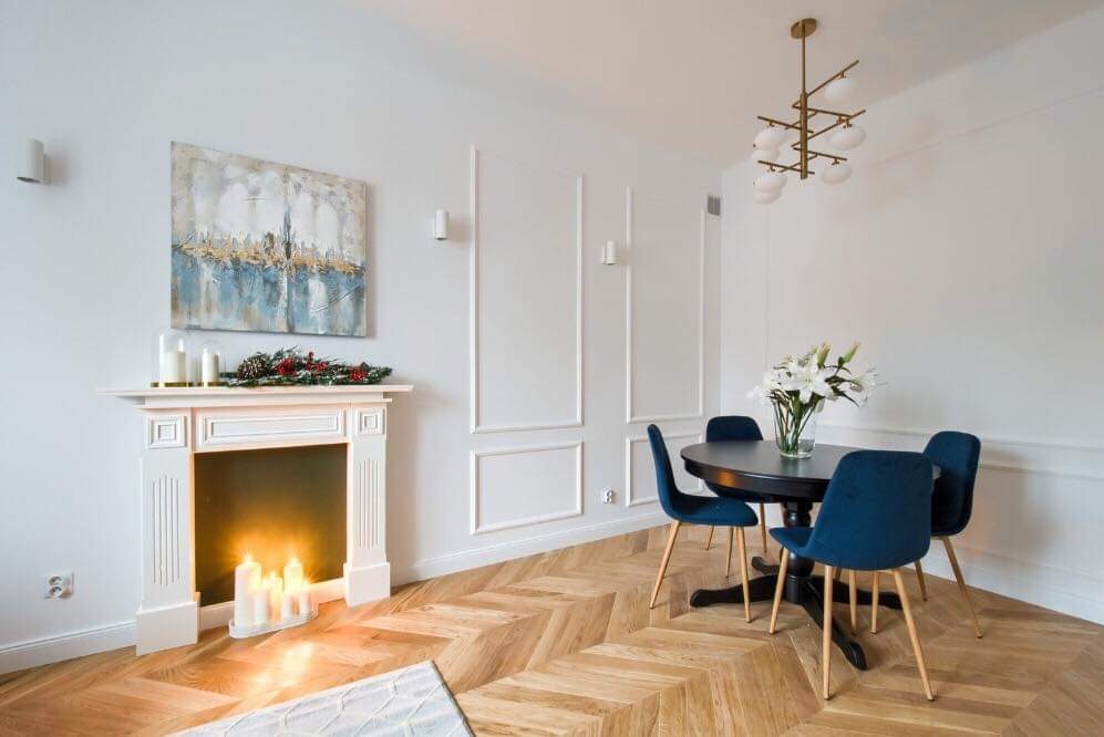 Jasne wnętrze z kominkiem, nad którym wisi abstrakcyjny obraz; obok stół z ciemnym blatem i niebieskimi krzesłami na parkiecie ułożonym w jodełkę; w tle dekoracyjne listwy na ścianie