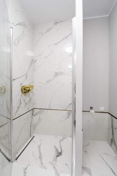 Nowoczesna kabina prysznicowa z białymi płytkami imitującymi marmur, złota armatura i czarne akcenty.