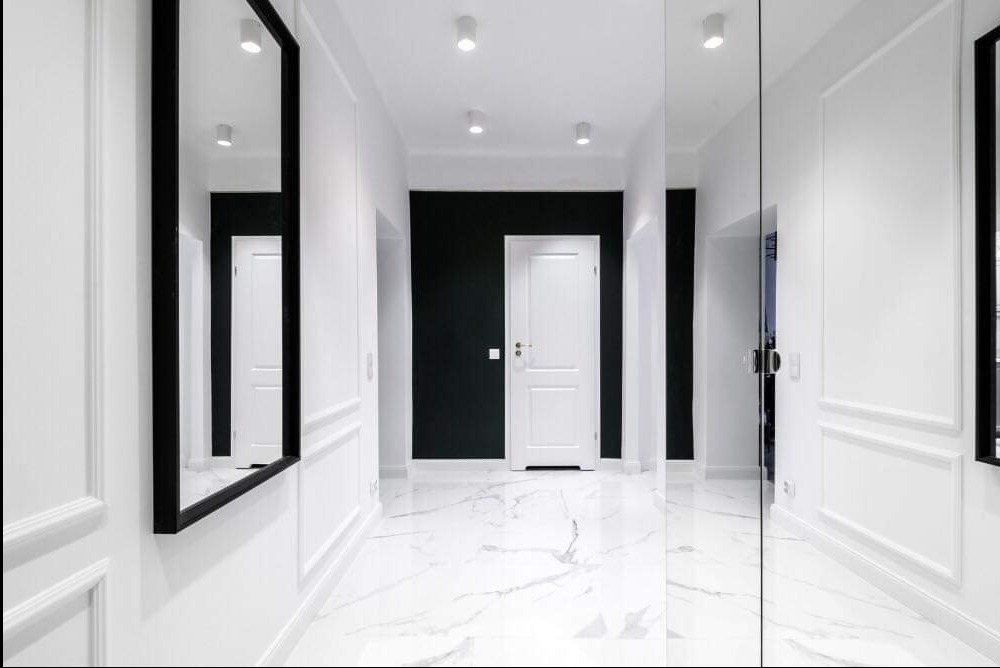 Biały korytarz z marmurową podłogą, czarną ścianą akcentującą i białymi drzwiami, ozdobne listwy ścienne, lustro w czarnej ramie.