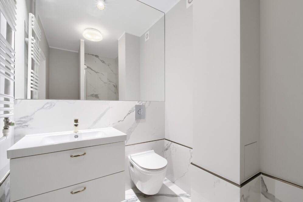 Nowoczesna, jasna łazienka z białymi marmurowymi płytkami, wiszącą toaletą i szafką umywalkową z złotymi uchwytami, odbicie kabiny prysznicowej w lustrze.