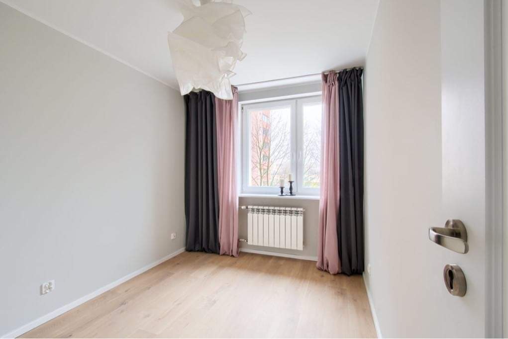 Jasne, puste pomieszczenie po odświeżeniu: parkiet w odcieniach beżu, szare ściany, okno z radiatorami, zasłony w kolorze pudrowego różu i grafitu, oraz nowoczesna, biała lampa wisząca.