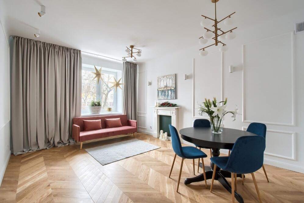 Jasne wnętrze salonu z parkietem w jodełkę, sofą w kolorze pudrowego różu, okrągłym stołem z bukietem lilii, dekoracyjnym kominkiem i minimalistycznym oświetleniem.