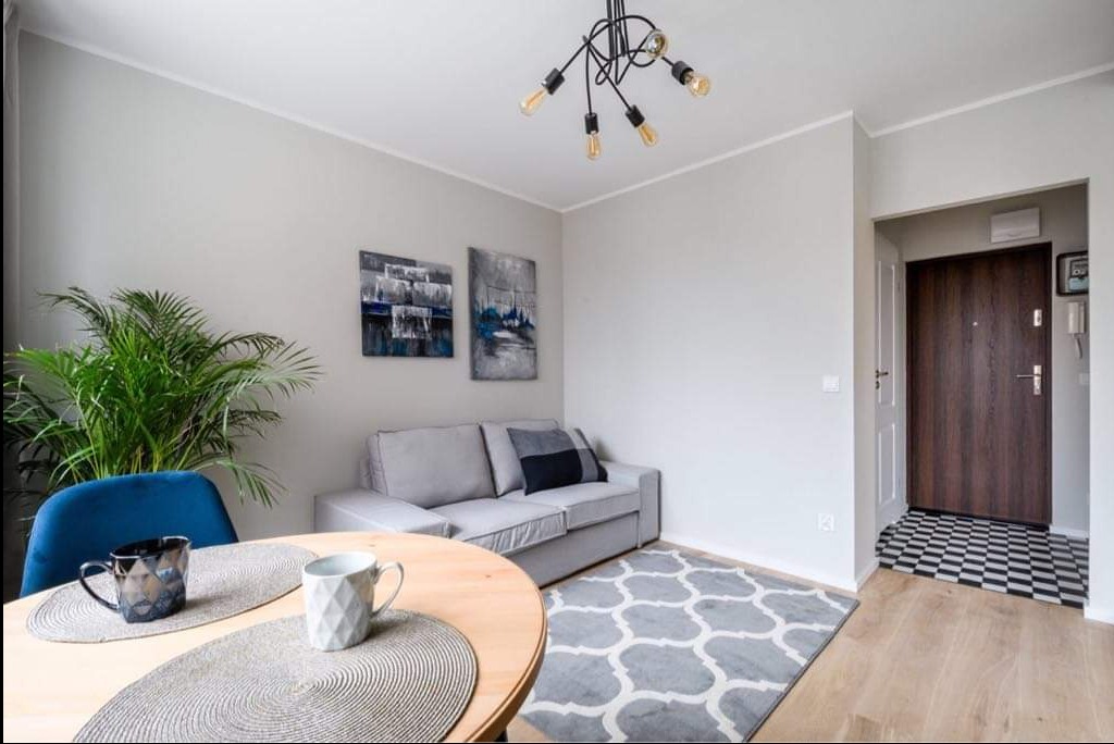 Jasne wnętrze mieszkania z drewnianym stołem, szarą sofą, dywanem w marokańskie wzory i nowoczesną lampą sufitową typu pająk.