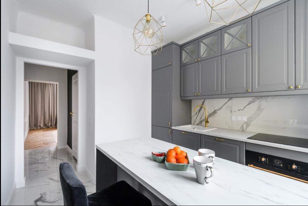 Nowoczesna kuchnia w odcieniach szarości z marmurowym blatem i złotymi akcentami, widok na przedpokój z zasłonami.