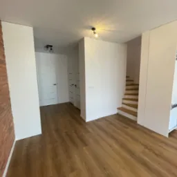 Jasne, przestronne wnętrze z drewnianą podłogą, ceglaną ścianą, białymi gładkimi ścianami i schodami z jasnego drewna.