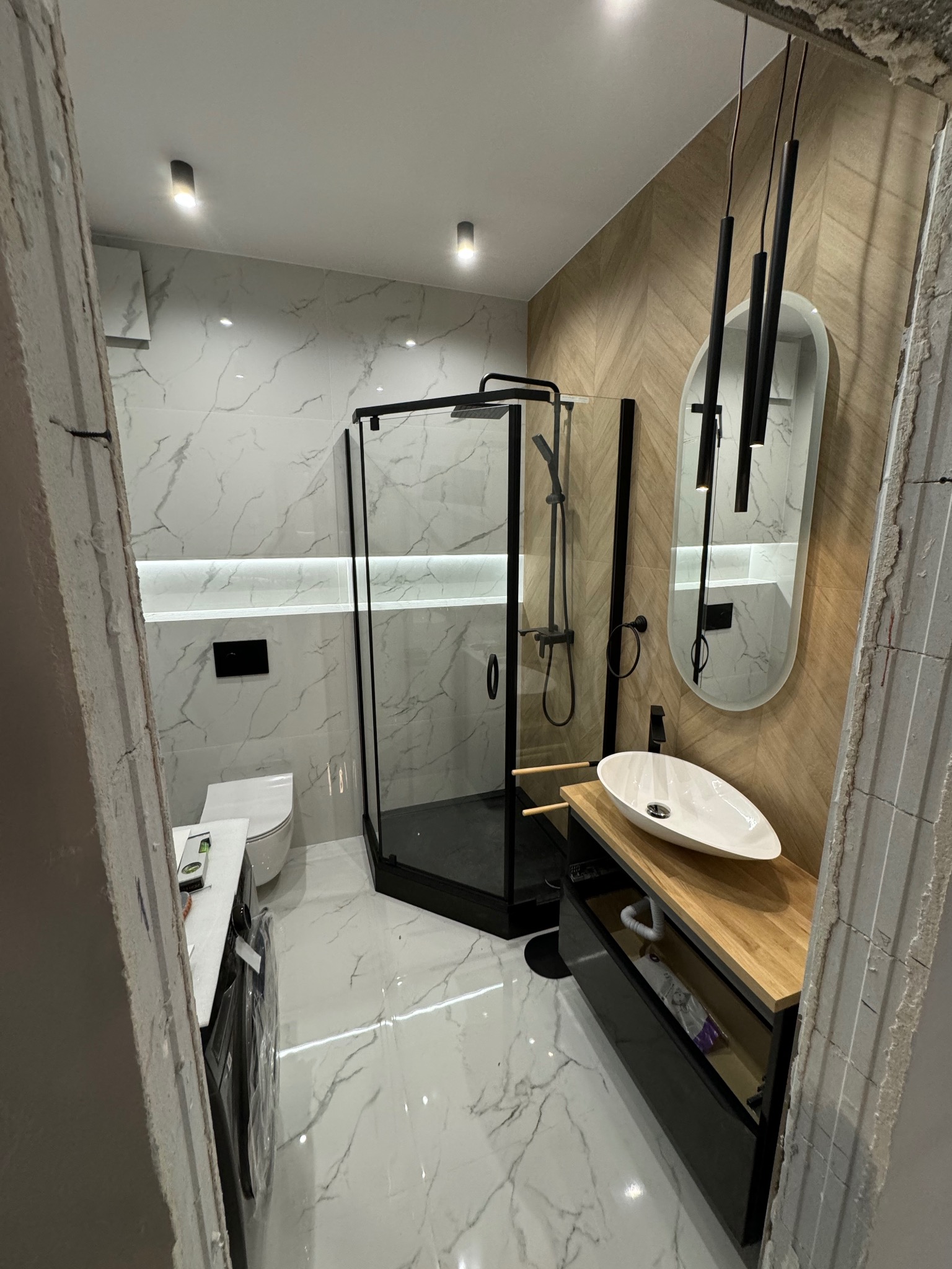 Nowoczesna łazienka w trakcie wykańczania: kabina prysznicowa z czarnymi profilami, umywalka nablatowa na drewnianym blacie, lustro owalne z wiszącymi lampami, płytki imitujące marmur na ścianach...
