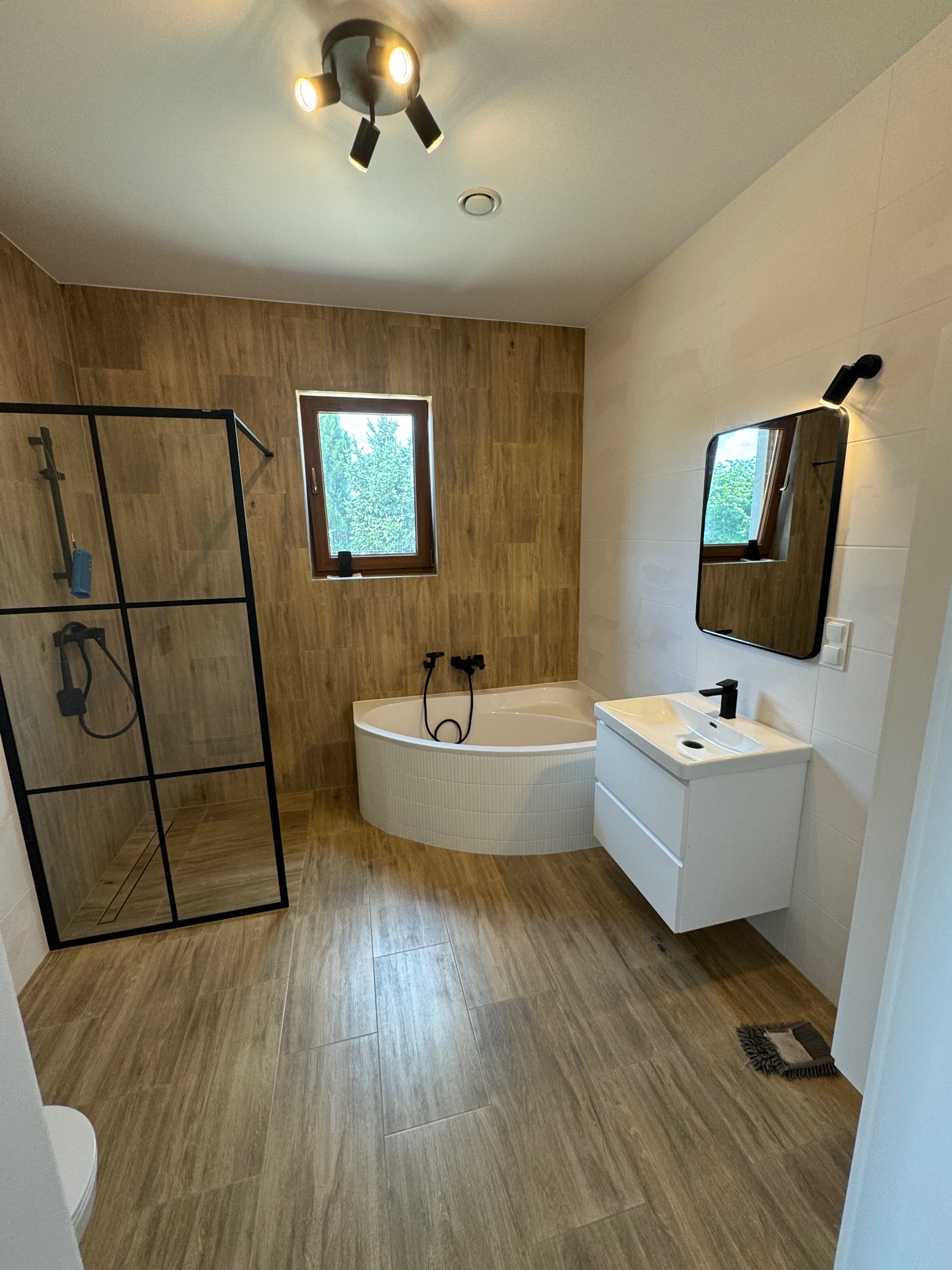 Nowoczesna łazienka z kabiną prysznicową typu walk-in, narożną wanną obudowaną mozaiką, umywalką z szafką i lustrem z podświetleniem LED, wykończona płytkami imitującymi drewno.