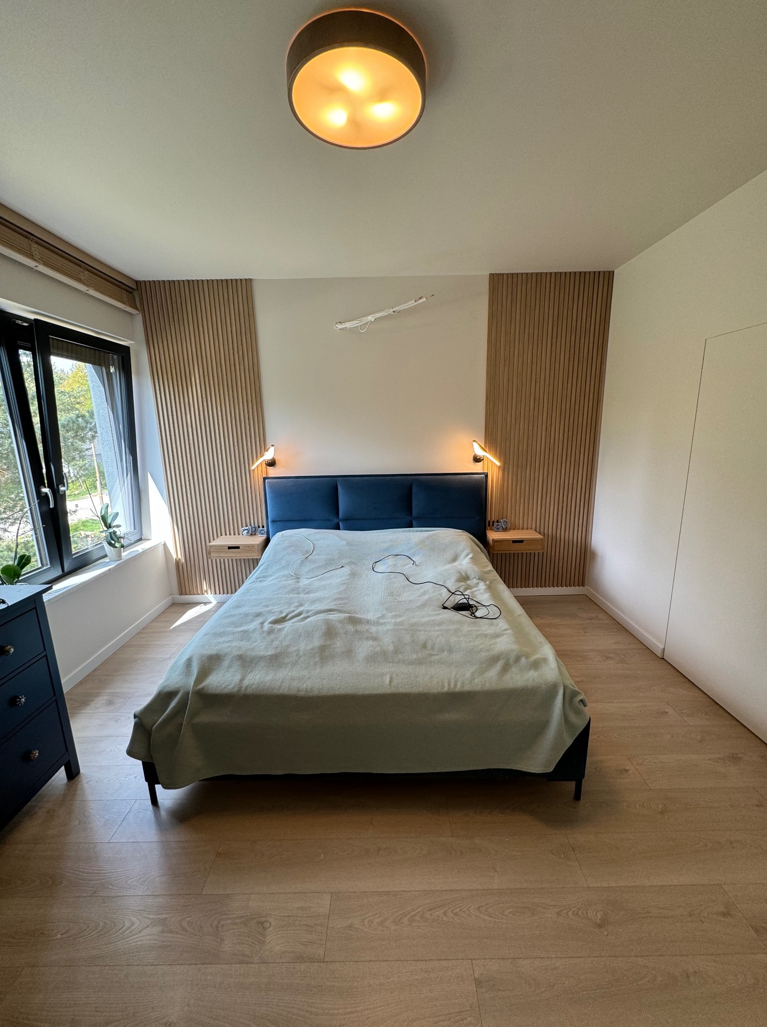 Nowoczesna sypialnia z drewnianymi panelami ściennymi po bokach łóżka z niebieskim wezgłowiem, drewnianą podłogą i szafkami nocnymi z lampkami.