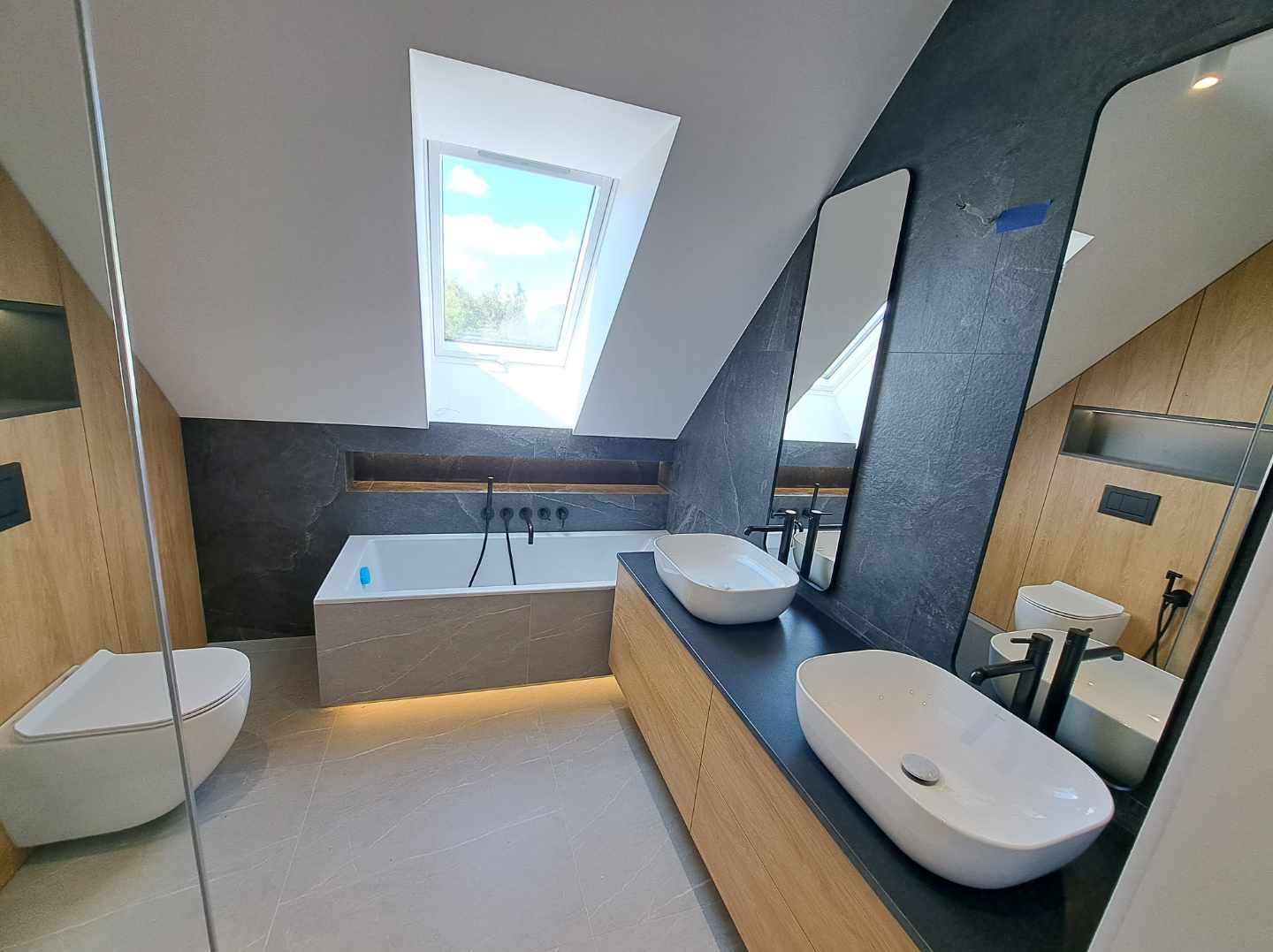 Nowoczesna łazienka na poddaszu z oknem dachowym, dwiema umywalkami nablatowymi, wanną z podświetleniem LED, szarą okładziną ścian i drewnianymi elementami wykończenia.