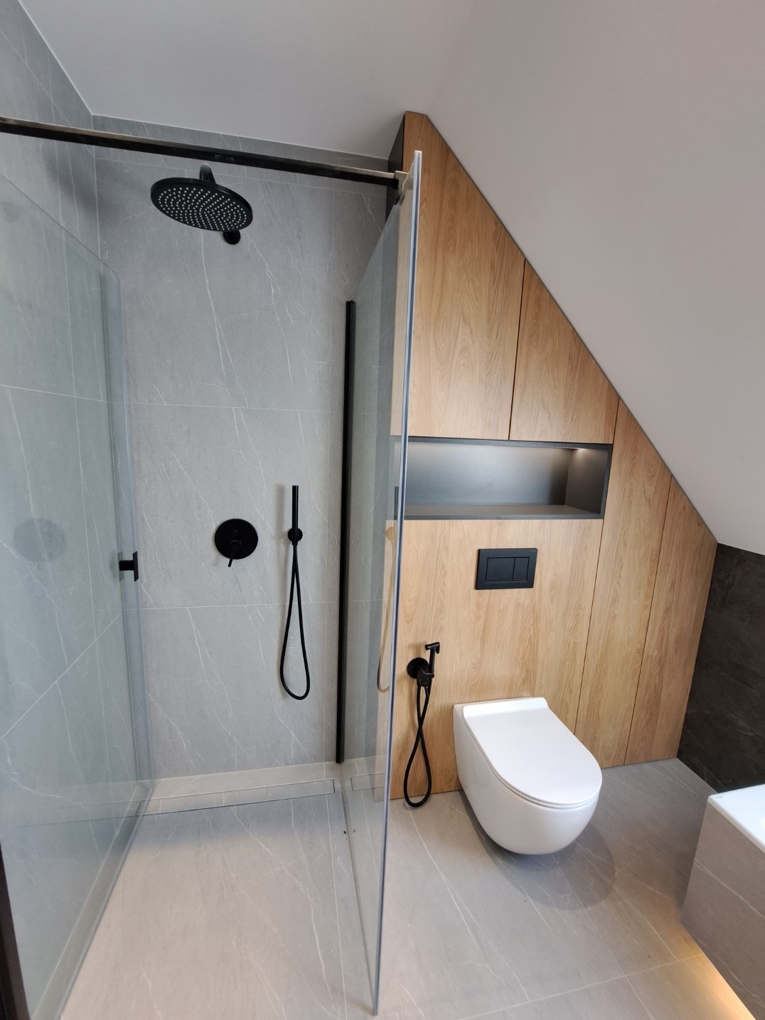 Nowoczesna łazienka na poddaszu z kabiną prysznicową walk-in, szarą płytką imitującą marmur, czarną armaturą i zabudową meblową z drewnopodobnej płyty.