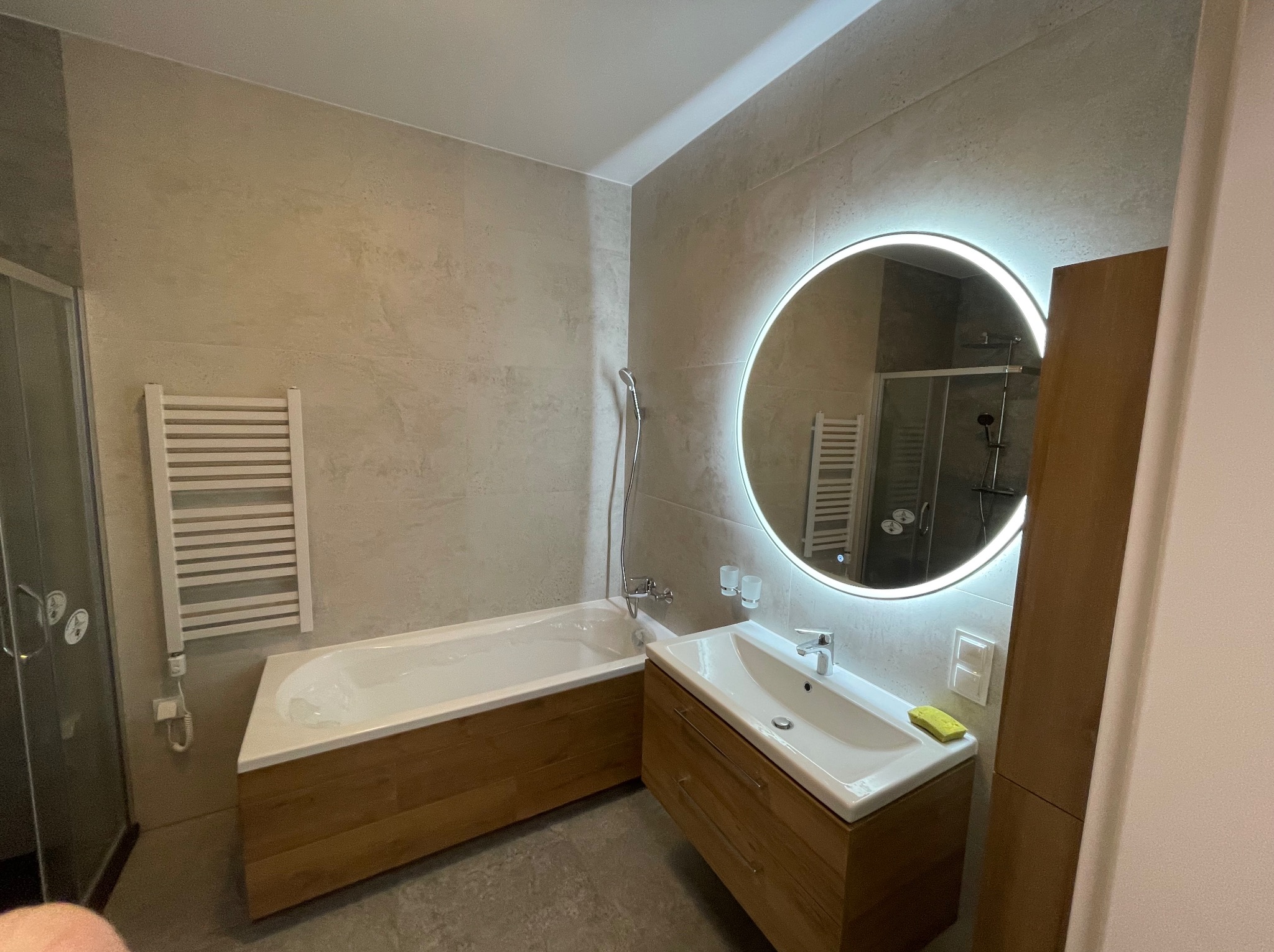 Nowoczesna łazienka z wanną w zabudowie z drewnianymi frontami, umywalką z szafką, okrągłym lustrem z podświetleniem LED i grzejnikiem drabinkowym na ścianie wykończonej płytkami w odcieniach beżu.