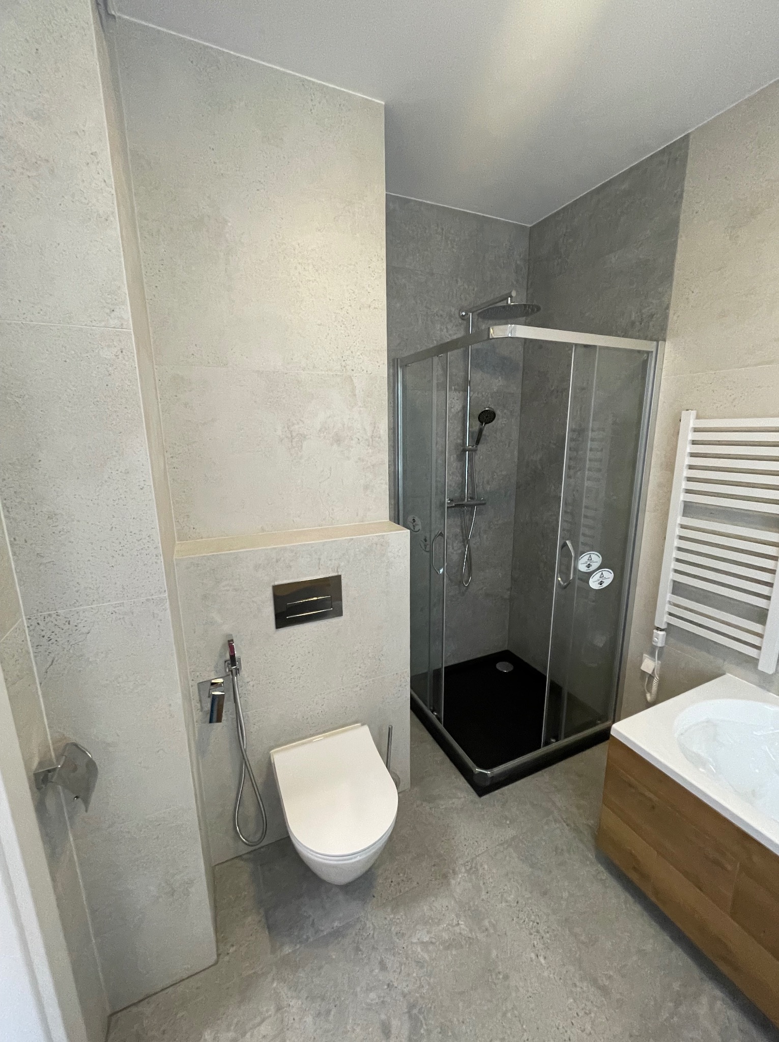 Nowoczesna łazienka z szarymi płytkami imitującymi kamień, kabiną prysznicową z czarnym brodzikiem, podwieszaną toaletą i drewnianą szafką pod umywalką.