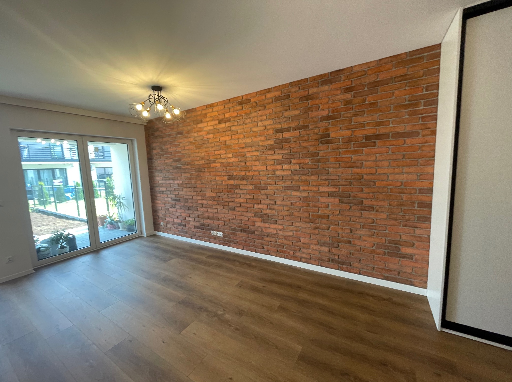 Pusty pokój z dekoracyjną ścianą z czerwonej cegły, panelami podłogowymi w kolorze brązowym, nowoczesną lampą sufitową i dużym oknem balkonowym.