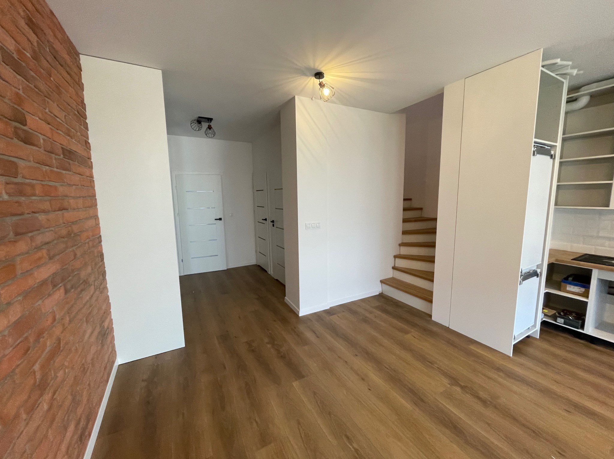 Jasne, przestronne wnętrze z drewnianą podłogą, ceglaną ścianą, białymi gładkimi ścianami i schodami z jasnego drewna.