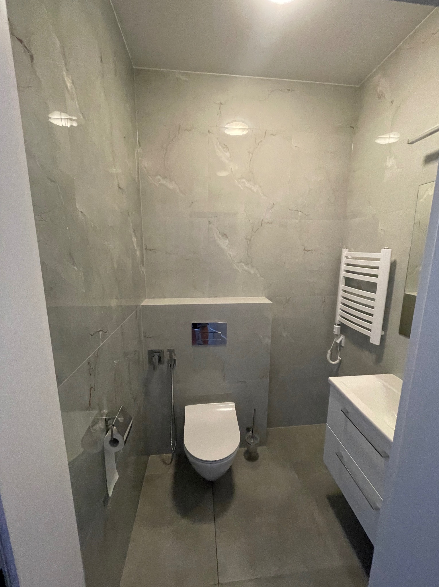 Nowoczesna łazienka z szarymi marmurkowymi płytkami, wiszącą toaletą, grzejnikiem drabinkowym i białą szafką z umywalką.