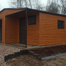 Zewnętrzny widok nowo postawionego, drewnianego garażu z bramą segmentową i drzwiami wejściowymi, otoczonego żwirem i rabatą z ziemią, na tle ogrodzenia i drzew bez liści.